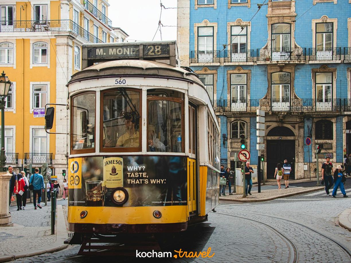 Żółty tramwaj 28 na brukowanej ulicy Lizbony z kolorowymi kamienicami.