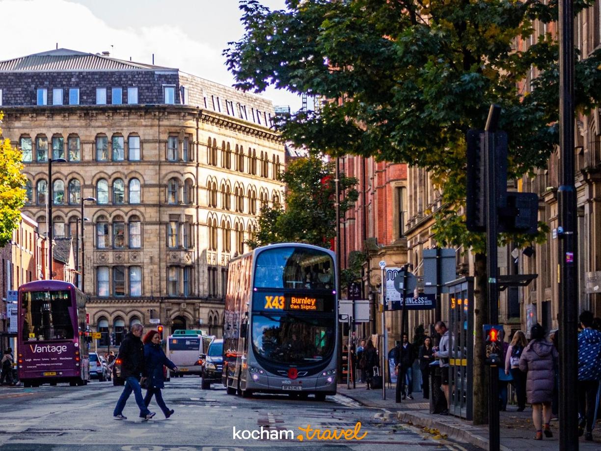 Jesienna ulica Manchesteru z piętrowym autobusem, ludźmi i zabytkowymi budynkami.