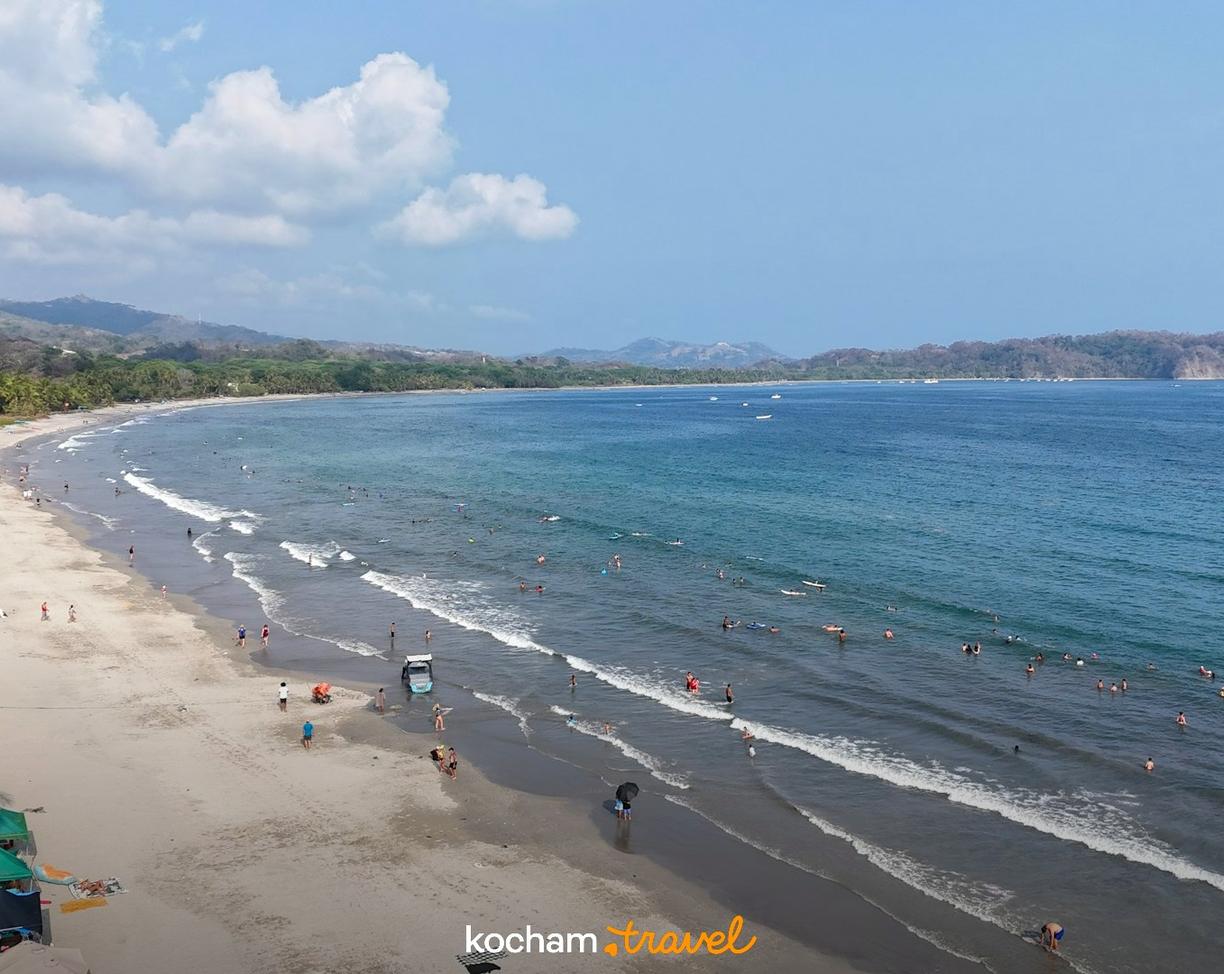Słoneczna plaża z kąpiącymi się ludźmi, palmami i lazurowym oceanem.