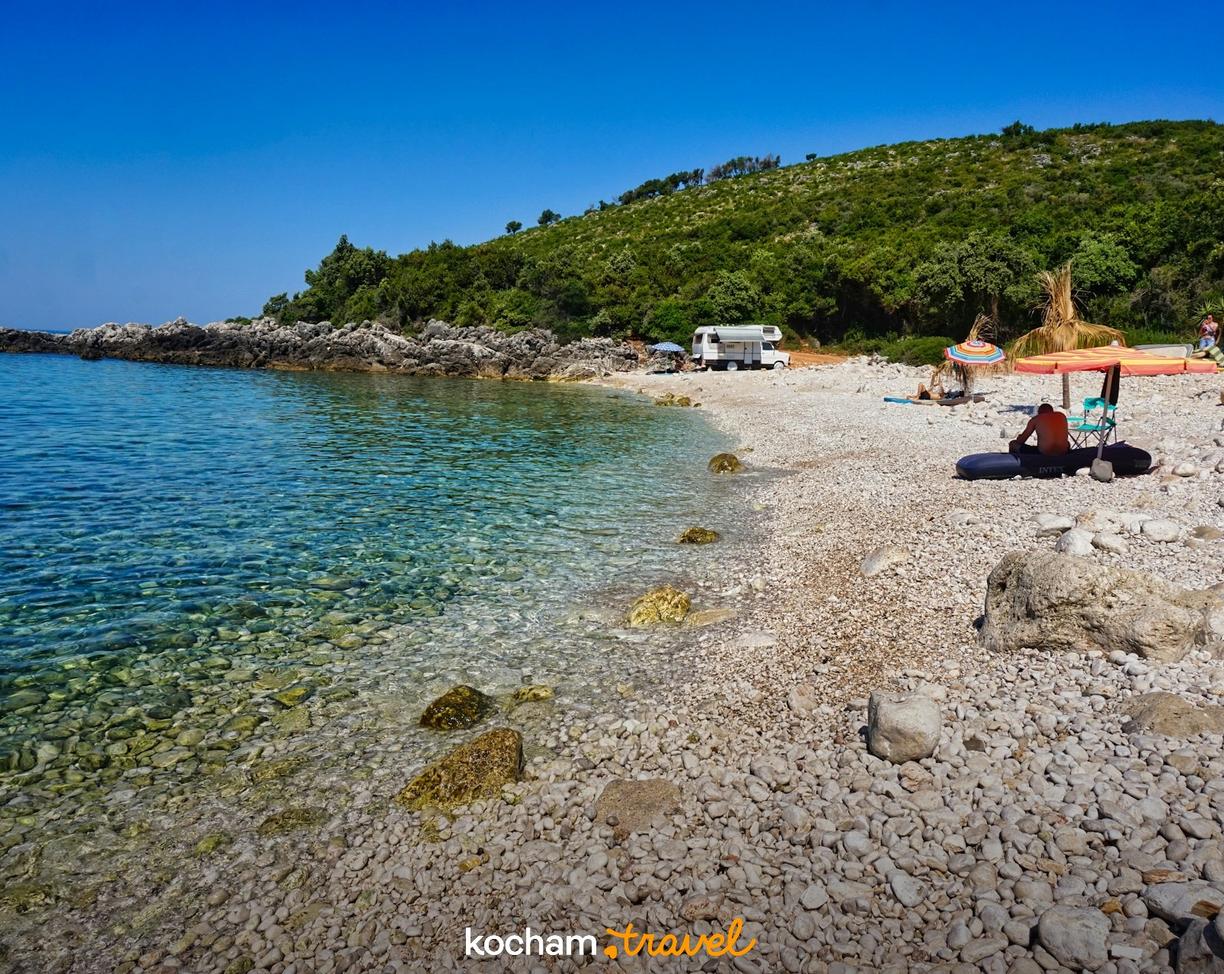 Malownicza kamienista plaża z krystaliczną wodą i kempingiem pod zielonymi wzgórzami.