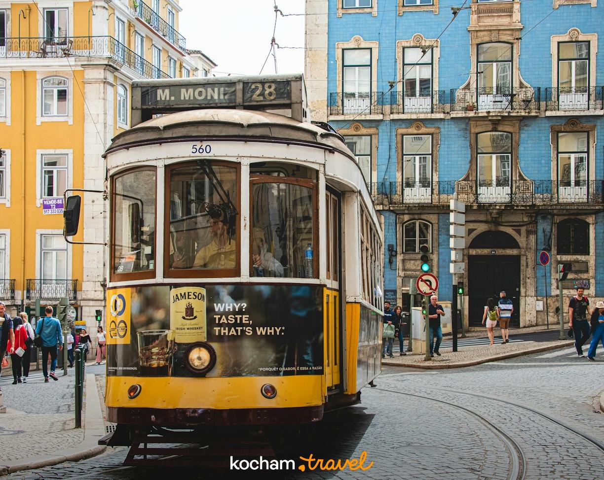 Żółty tramwaj 28 na brukowanej ulicy Lizbony z kolorowymi kamienicami.