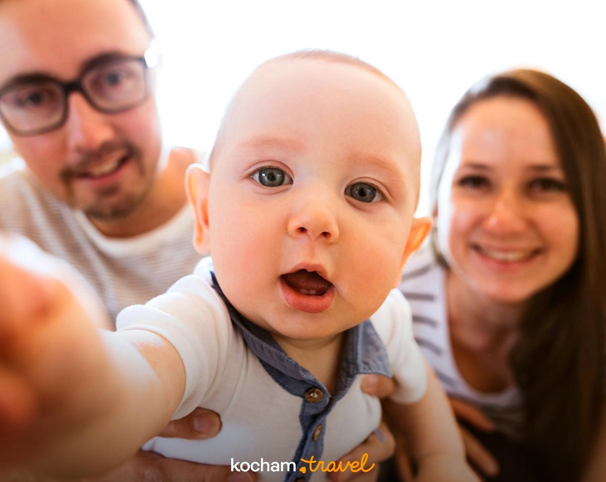 Niemowlę robiące selfie z uśmiechniętymi rodzicami w tle.
