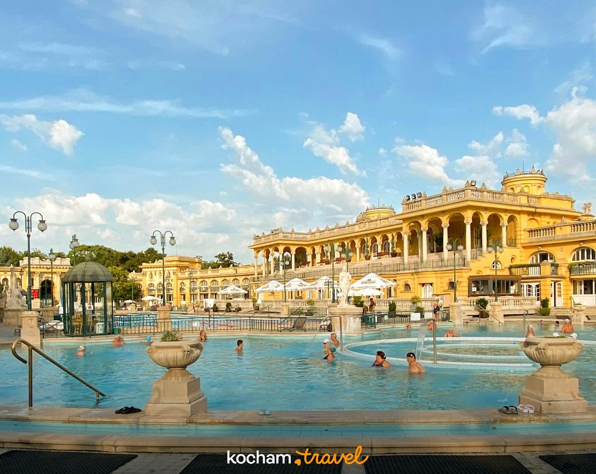 Kąpielisko termalne Szechenyi w Budapeszcie, żółte budynki, relaksujący się ludzie.
