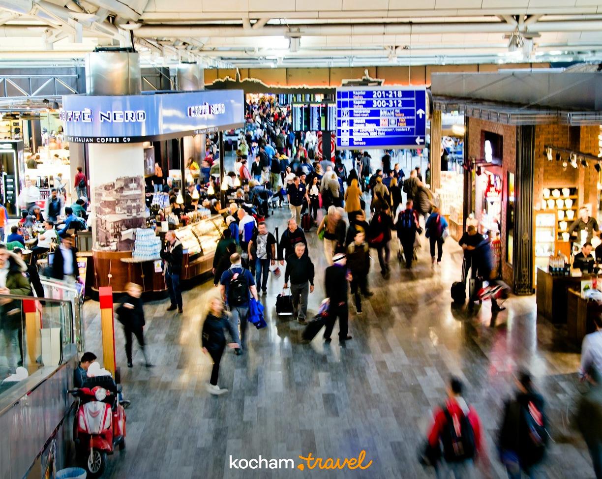 Zatłoczony terminal lotniska z podróżnymi, sklepami i tablicą informacyjną.
