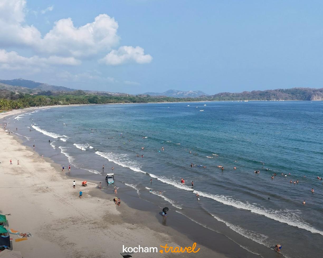 Słoneczna plaża z kąpiącymi się ludźmi, palmami i lazurowym oceanem.