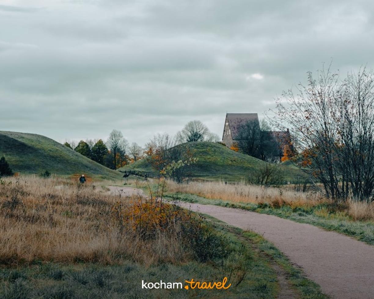 Ścieżka, zielone kurhany, historyczny budynek, pochmurne jesienne niebo.