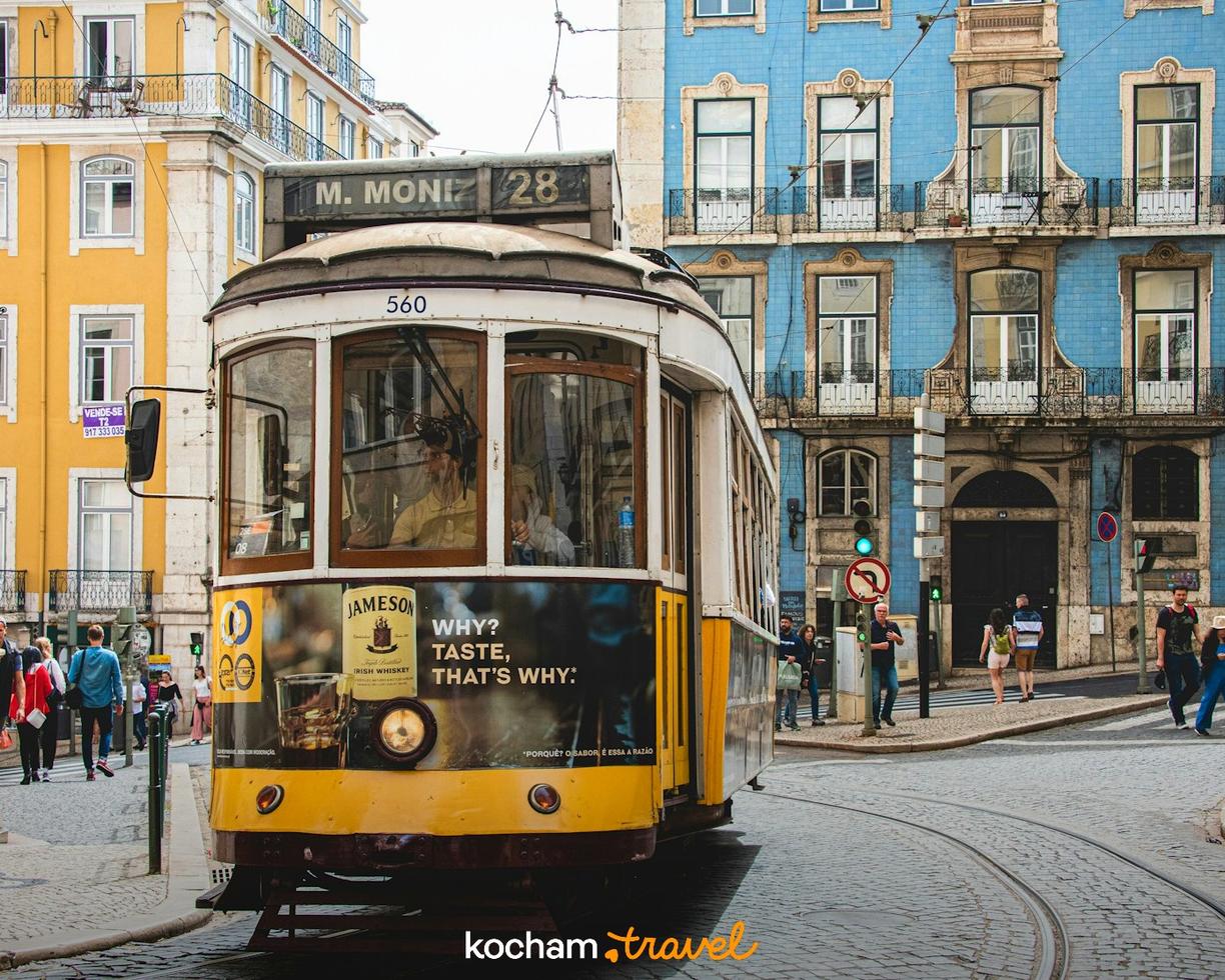 Żółty tramwaj 28 na brukowanej ulicy Lizbony z kolorowymi kamienicami.