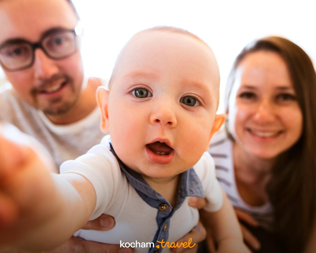 Niemowlę robiące selfie z uśmiechniętymi rodzicami w tle.