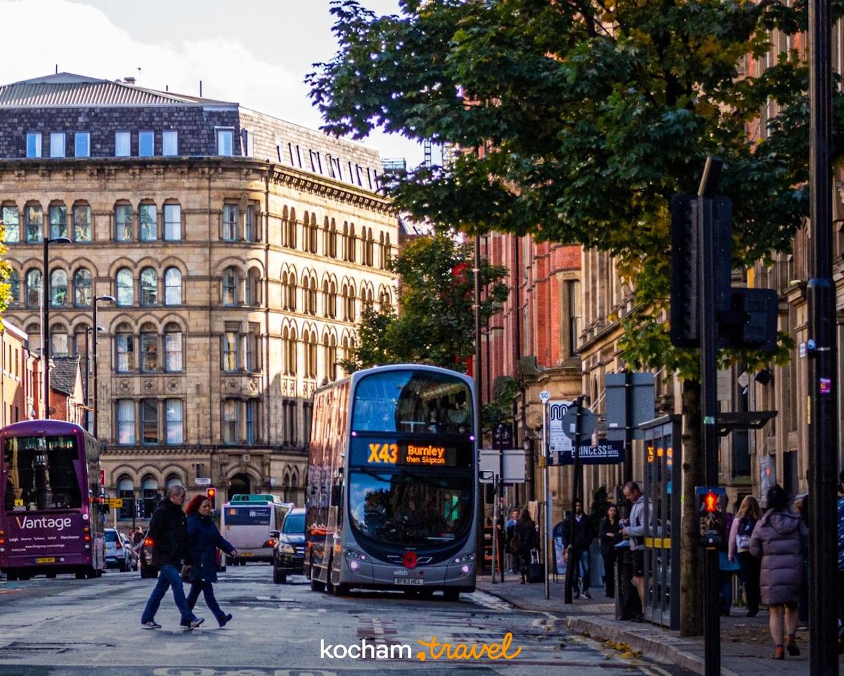 Jesienna ulica Manchesteru z piętrowym autobusem, ludźmi i zabytkowymi budynkami.