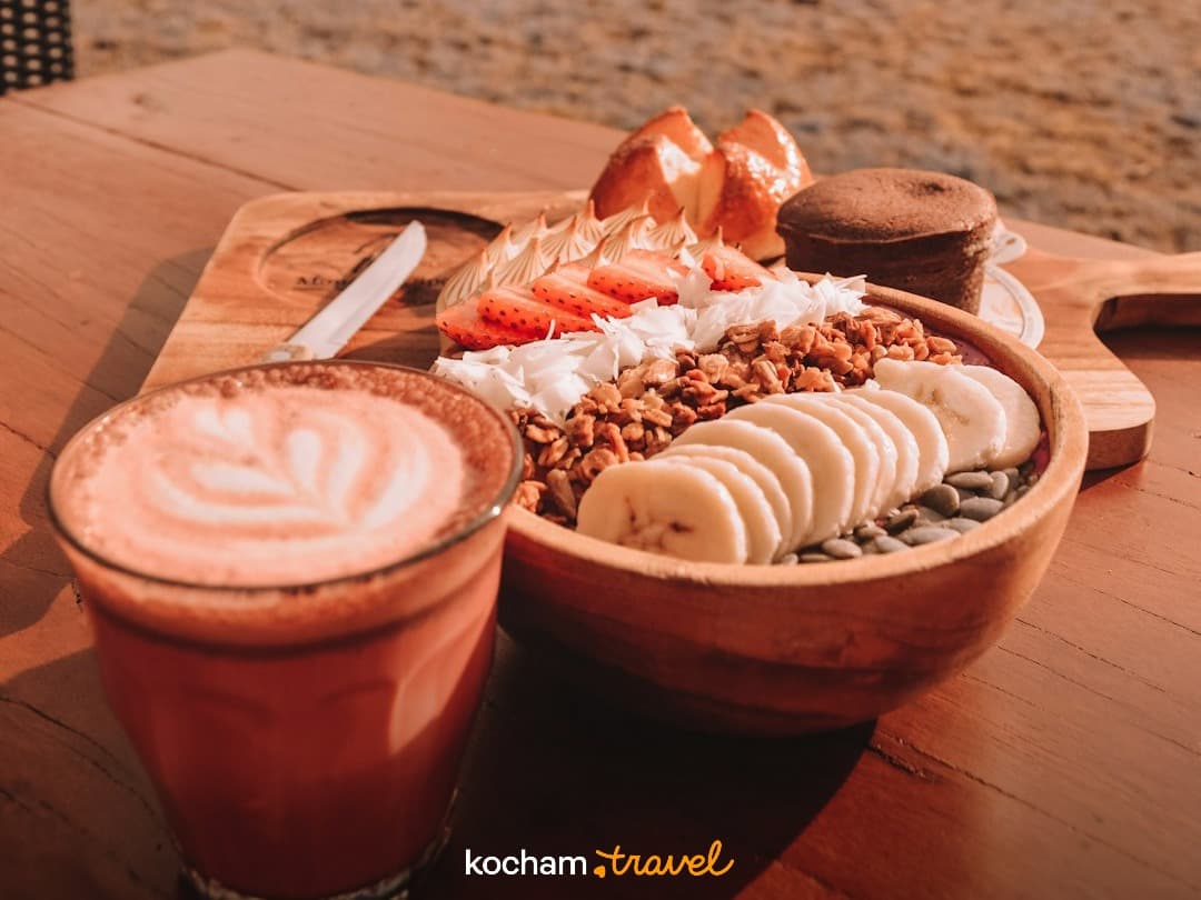 Śniadaniowy stół: miska acai, kawa latte, owoce i granola na drewnie.