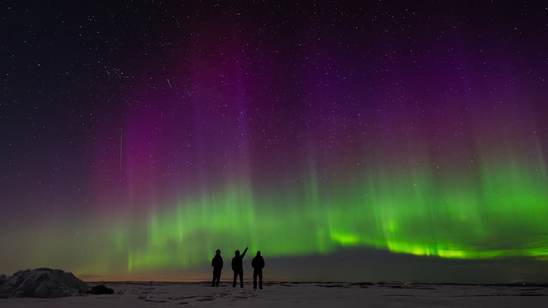 Spektakularna zielono-fioletowa zorza polarna i sylwetki ludzi na śniegu.