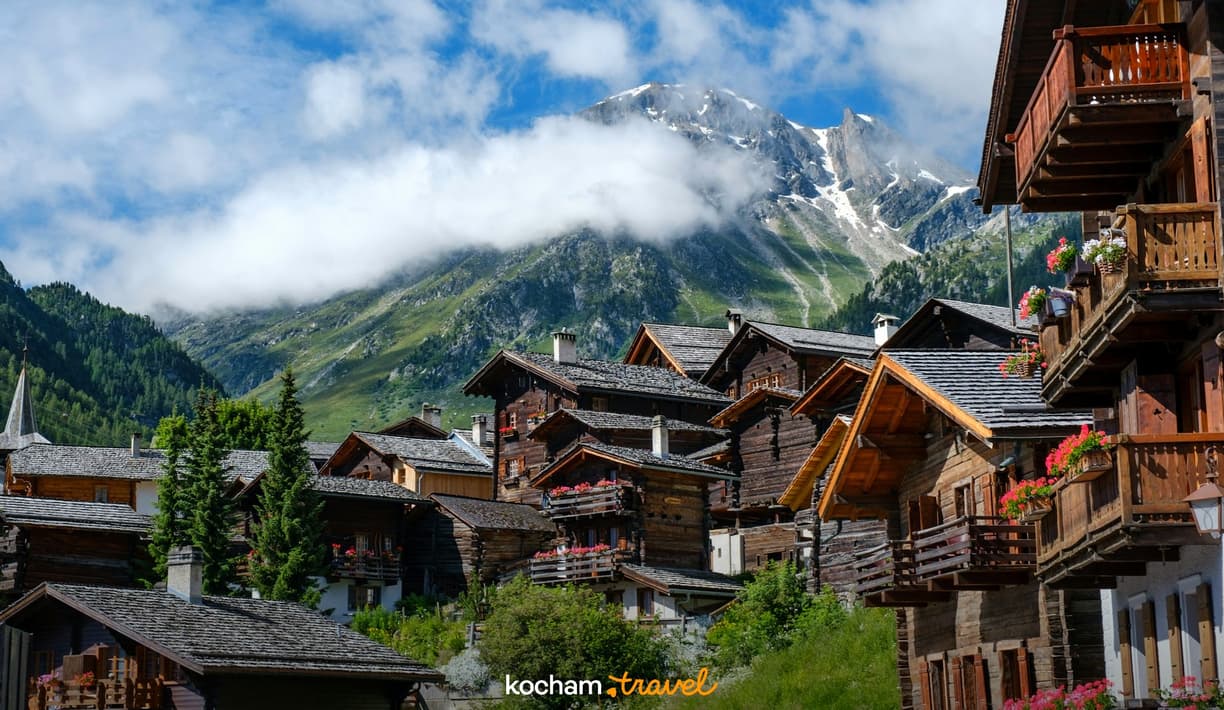 Alpejska wioska z drewnianymi domami, kwitnącymi balkonami i ośnieżonymi górami.