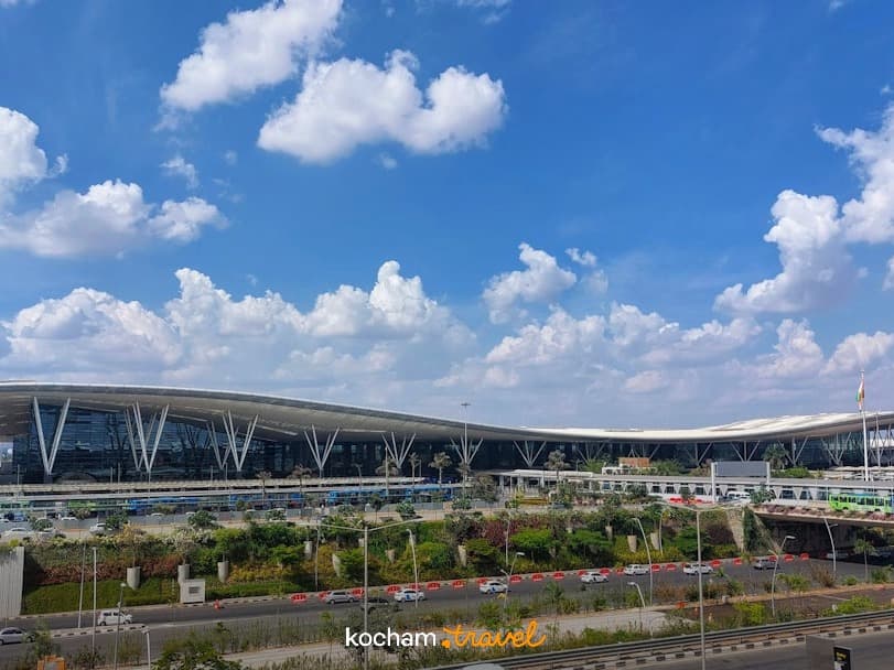 Nowoczesny terminal lotniska z falistym dachem, jasne niebo, ruch samochodowy.