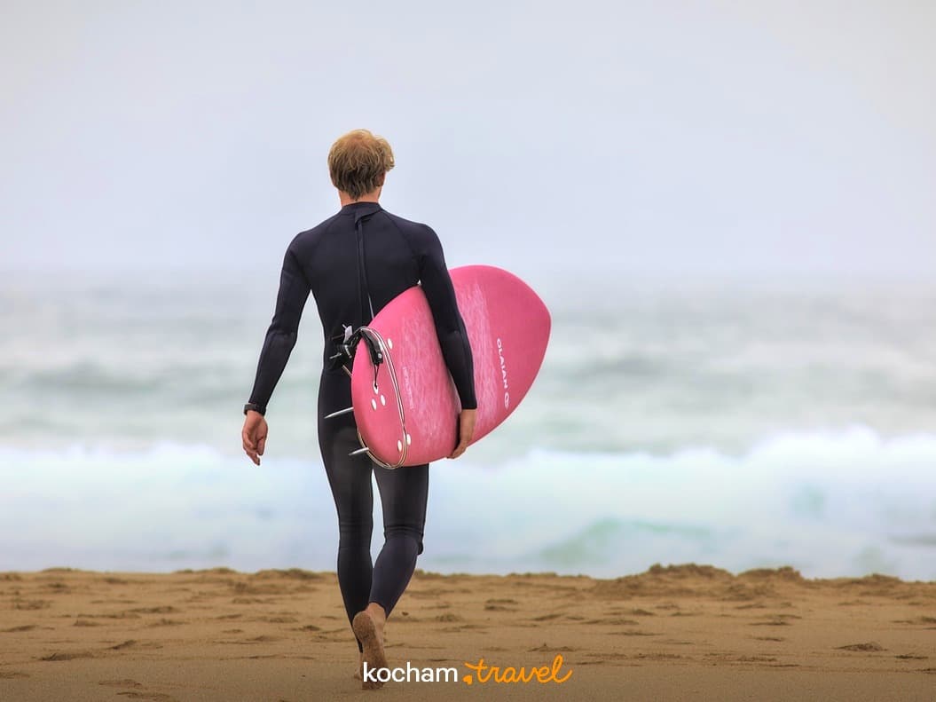 Surfer w czarnym kombinezonie z różową deską idzie na plaży.