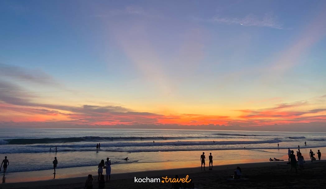Plaża o zachodzie słońca, ludzie, surferzy, kolorowe niebo i księżyc.