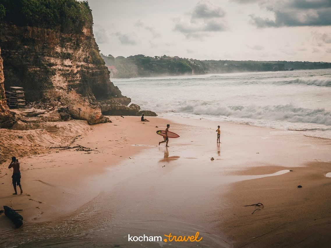 Surfer niosący deskę na rozległej piaszczystej plaży z klifami i falami oceanu.