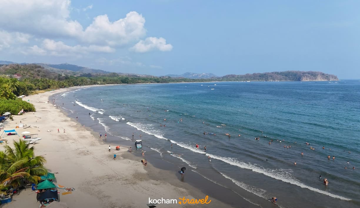Słoneczna plaża z kąpiącymi się ludźmi, palmami i lazurowym oceanem.