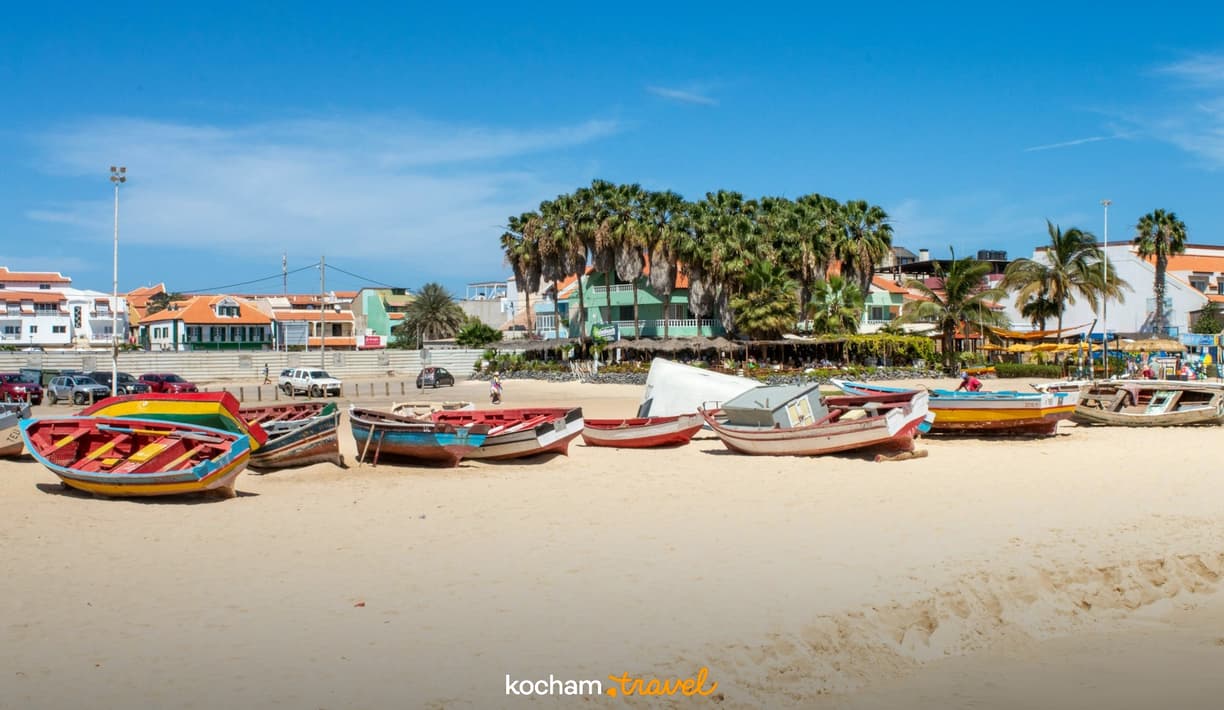 Piaszczysta plaża z kolorowymi łodziami i tropikalnym miasteczkiem w tle.