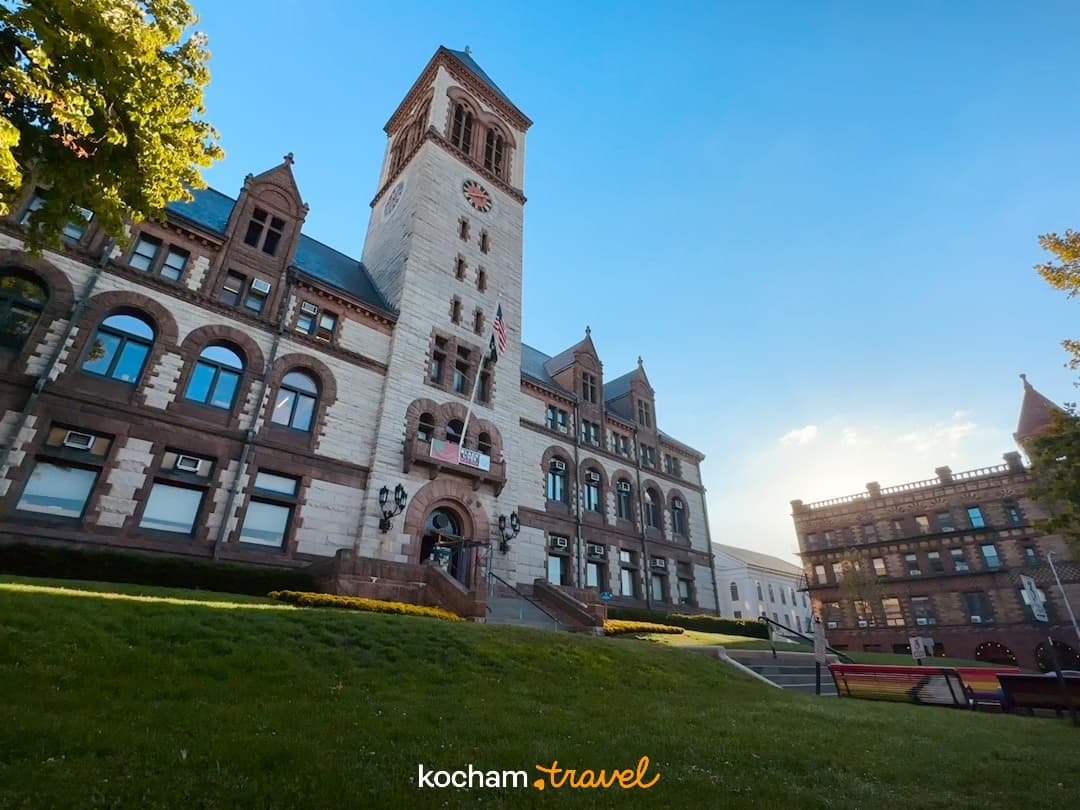 Historyczny kamienny budynek z wieżą zegarową, słoneczny dzień, zielona trawa, błękitne niebo.