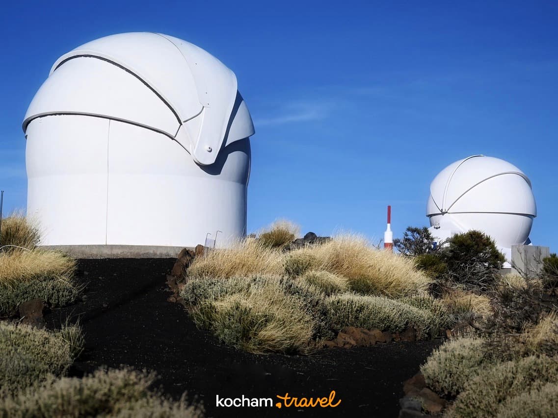 Białe kopuły obserwatorium astronomicznego na wulkanicznym krajobrazie pod błękitnym niebem.