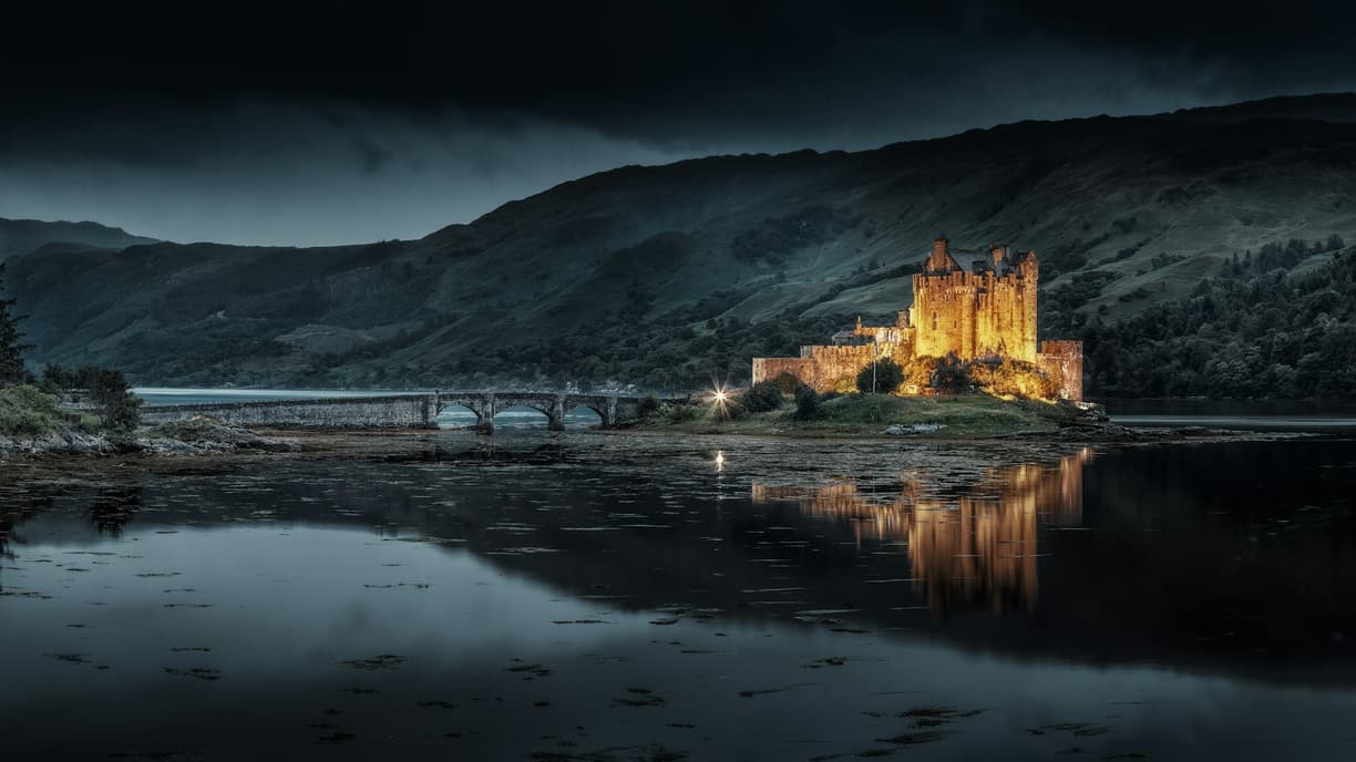 Zamek Eilean Donan oświetlony nocą, z mostem i odbiciem w wodzie.
