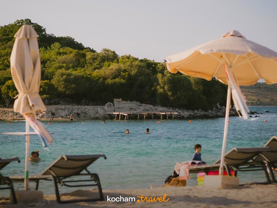 Słoneczna plaża z turkusową wodą, ludźmi, parasolami i zielonymi wzgórzami.