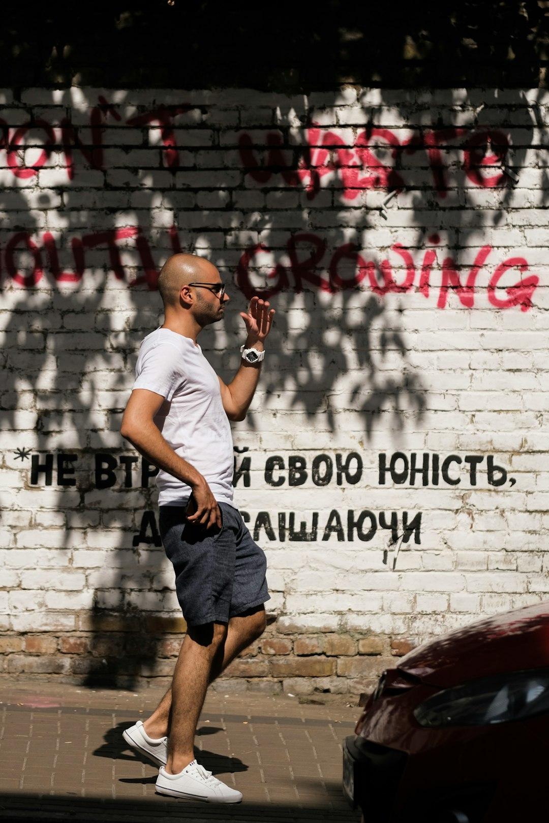 Łysy mężczyzna w okularach i białym t-shircie, spacerujący przy ścianie z graffiti.
