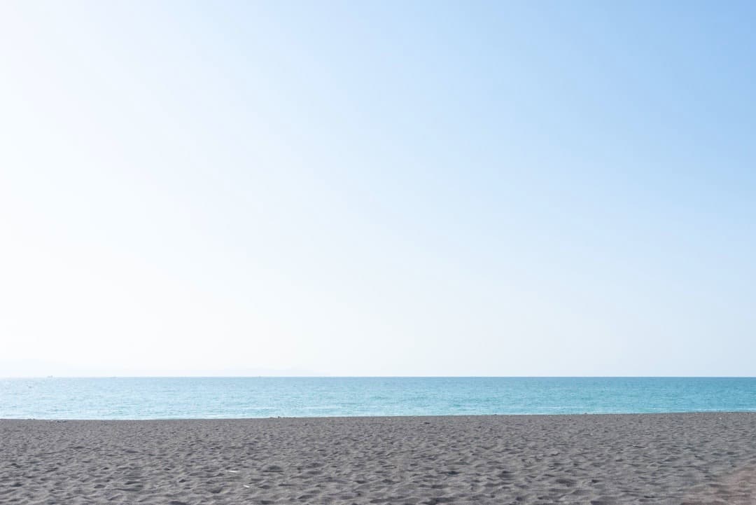 Słoneczny dzień na plaży z ciemnym piaskiem i turkusową wodą.