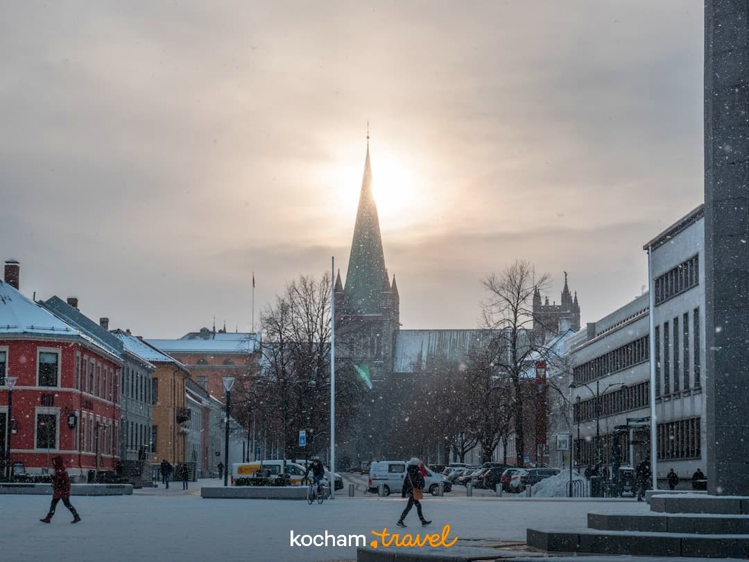 Zasnieżony plac miejski z Katedrą Nidaros, padający śnieg, słońce.