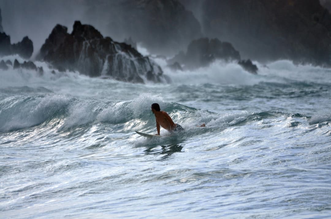 Surfer na desce płynący przez wzburzone fale oceanu, skały w tle.