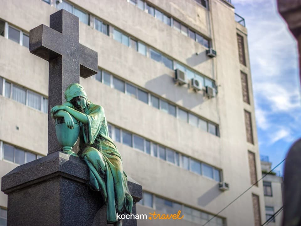 Brązowa statua żałobnej figury, krzyż, cmentarz miejski, budynek.