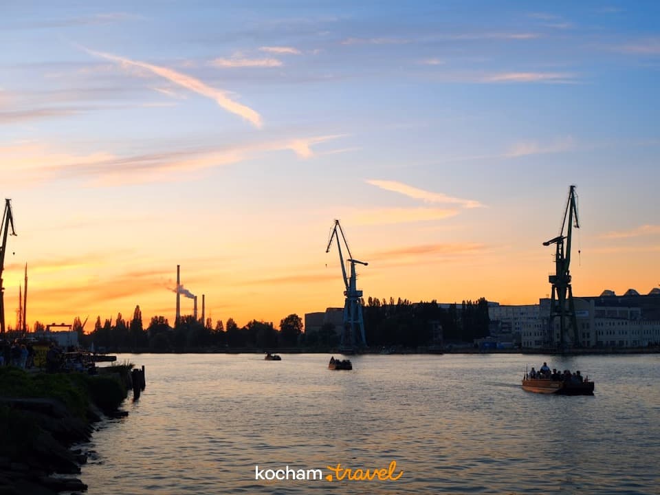 Rzeczny port o zachodzie słońca z żurawiami przemysłowymi i łodziami.