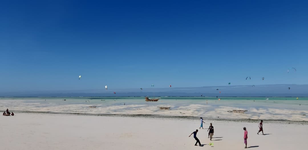 Plaża z kitesurferami, ludźmi grającymi w piłkę i łodziami.