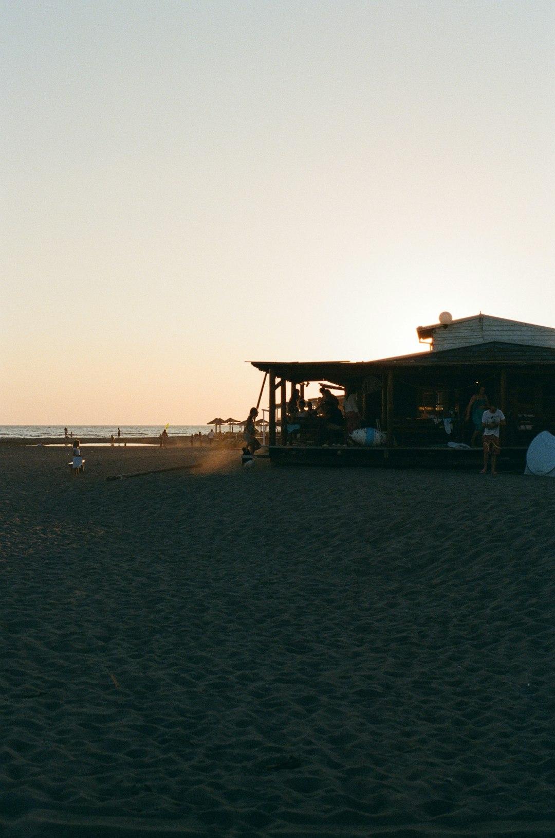 Plaża o zachodzie słońca z barem, ludźmi i ciemnym piaskiem.