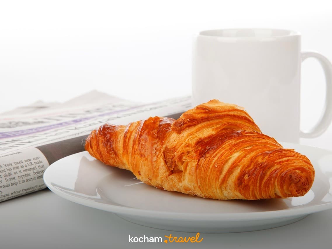 Chrupiący croissant na talerzu, biała filiżanka kawy i gazeta na śniadanie.