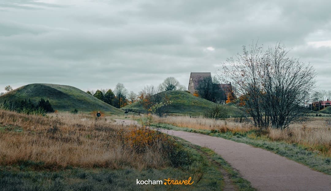 Ścieżka, zielone kurhany, historyczny budynek, pochmurne jesienne niebo.