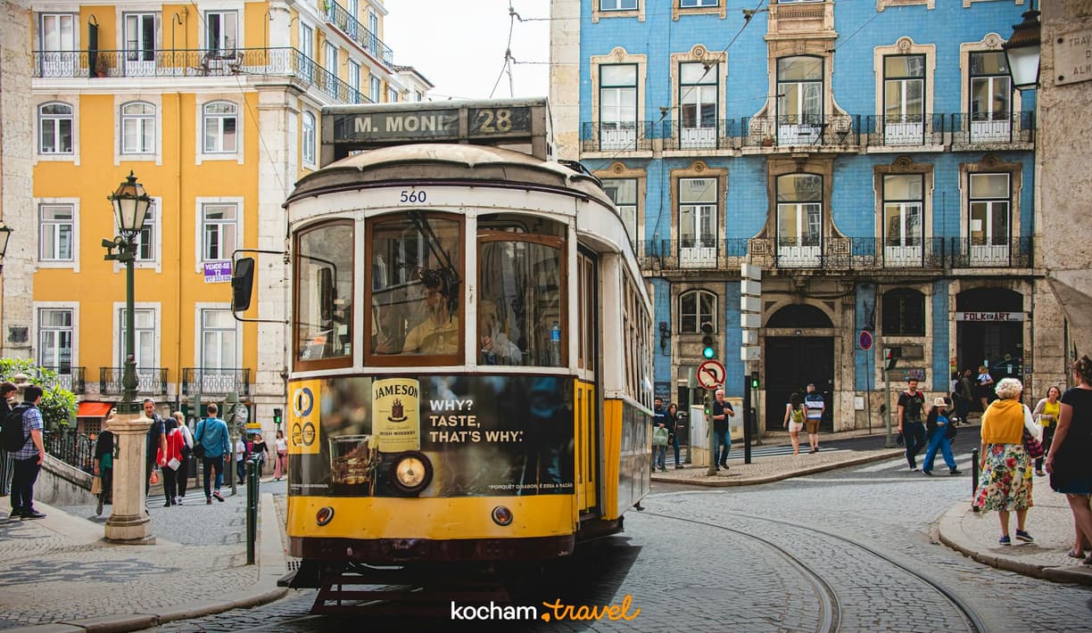 Żółty tramwaj 28 na brukowanej ulicy Lizbony z kolorowymi kamienicami.