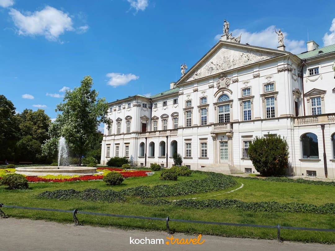 Majestatyczny biały pałac, zielony ogród, fontanna, słoneczny dzień.