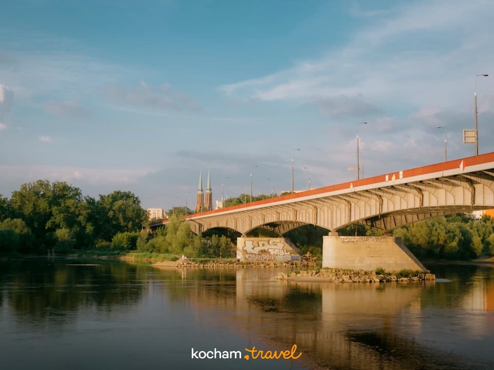 Most nad rzeką, zielone brzegi, miejska panorama, zachód słońca.