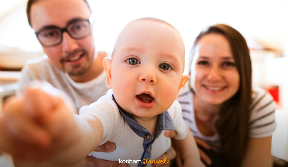 Niemowlę robiące selfie z uśmiechniętymi rodzicami w tle.