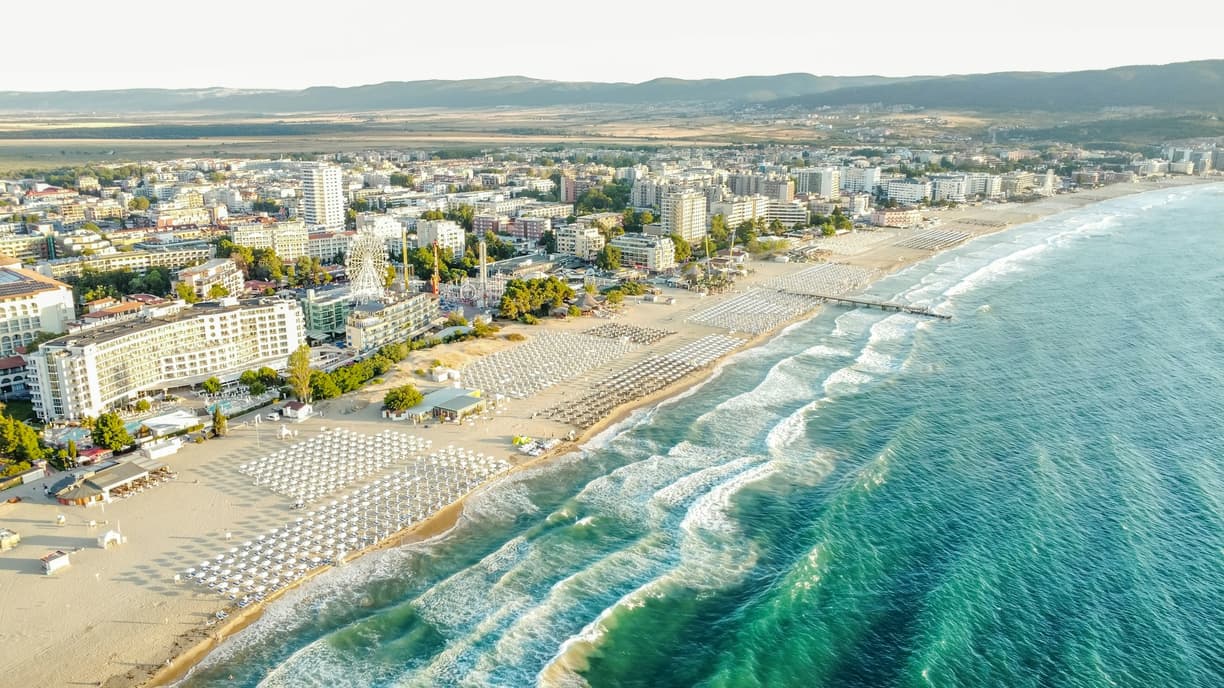 Piaszczysta plaża z rzędami parasoli, hotele i turkusowe morze.