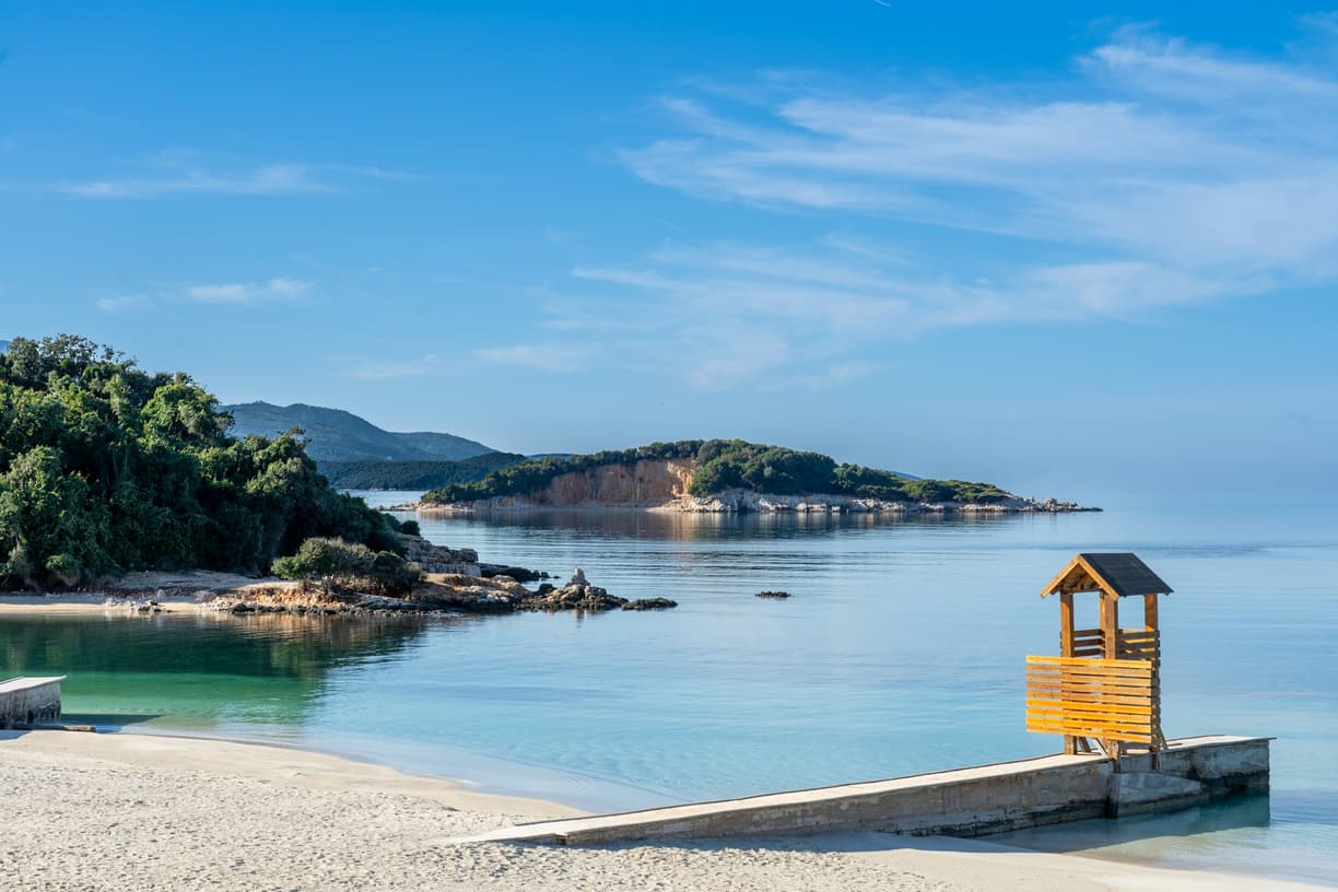 Spokojna zatoka z piaszczystą plażą, turkusową wodą i drewnianą wieżą.