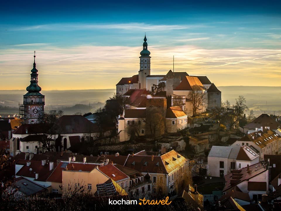 Panorama europejskiego miasta z zamkiem i dwiema wieżami o zachodzie słońca.