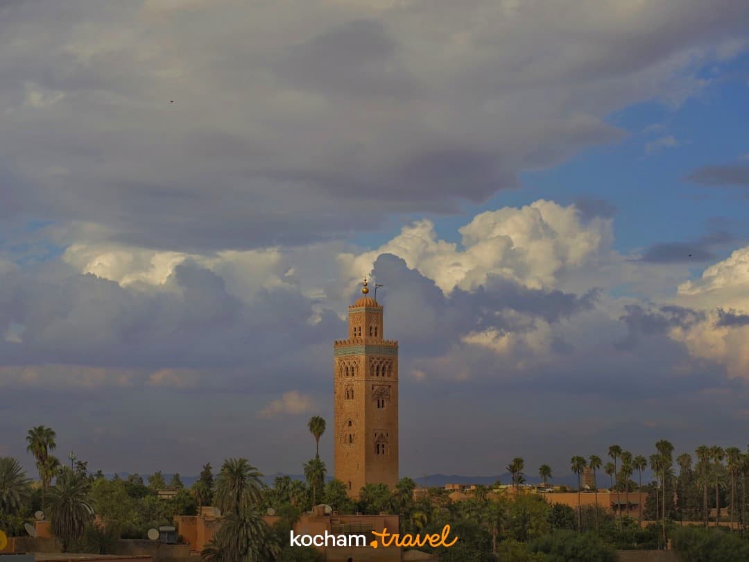 Wysoki minaret, palmy, budynki, dramatyczne niebo nad miastem.
