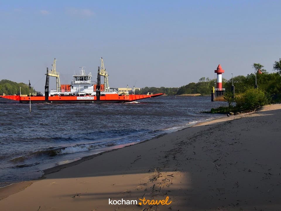 Prom JULIUSPLATE, rzeka, piaszczysta plaża, latarnia morska, słoneczny dzień.