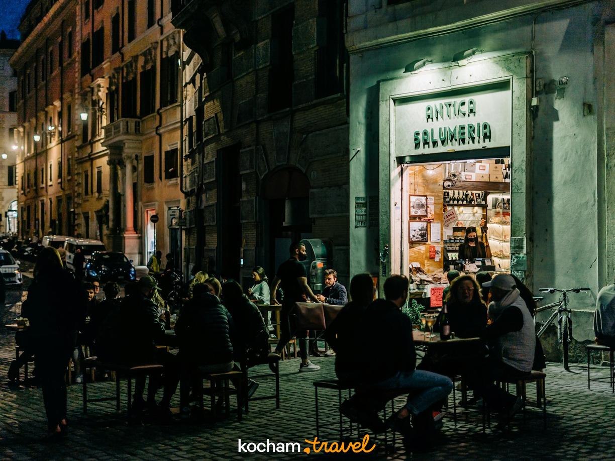 Goście jedzą i rozmawiają przy stolikach przed Antica Salumeria nocą.