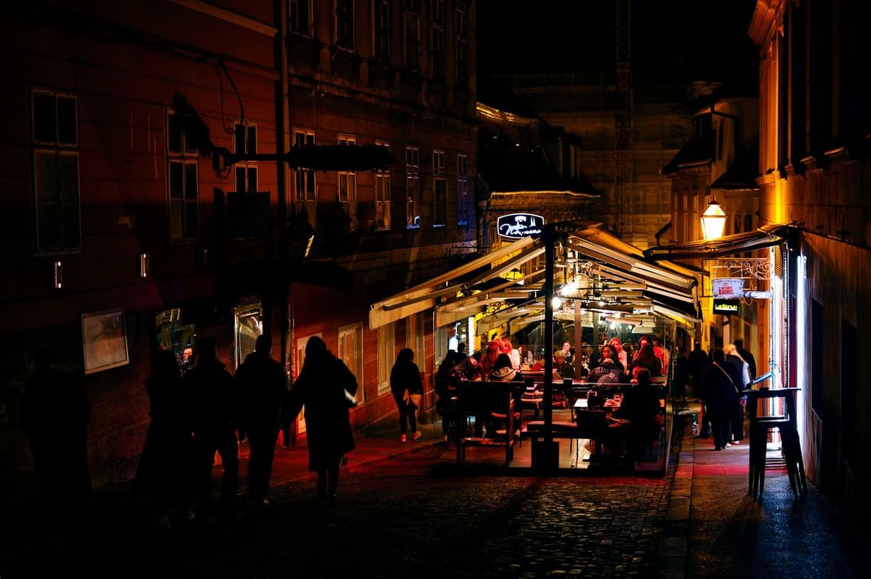 Tętniąca życiem ulica nocą z oświetlonym tarasem restauracji i przechodniami.