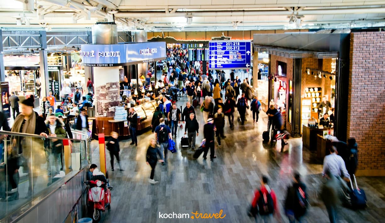 Zatłoczony terminal lotniska z podróżnymi, sklepami i tablicą informacyjną.