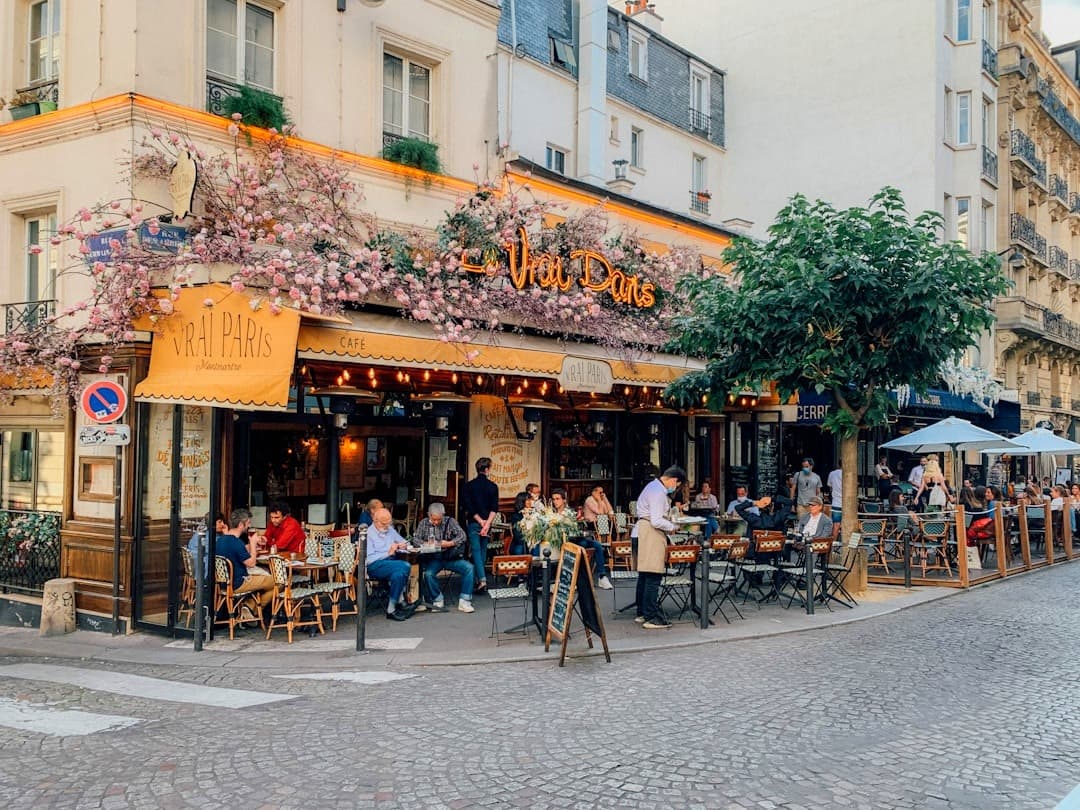 Paryska kawiarnia Vrai Paris z różowymi kwiatami, ludźmi przy stolikach na ulicy.