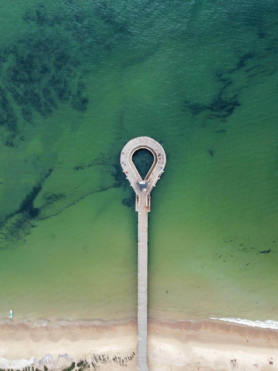 Molo w kształcie pętli, czyste zielonkawo-niebieskie morze, piaszczysta plaża z góry.